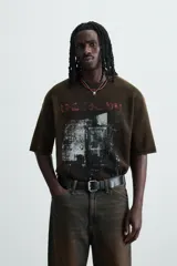 Camiseta de punto de algodón con corte boxy fit, cuello redondo y manga corta. Presenta un estampado frontal con efecto desgastado en tonos blanco y rojo sobre un fondo marrón oscuro/verde oliva lavado.