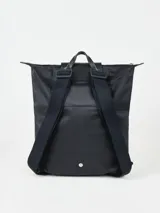Mochila de diseño minimalista en color beige, con cierre superior de cremallera y solapa frontal con botón a presión. Presenta un asa superior y correas ajustables, con el logo de la marca grabado en la solapa.