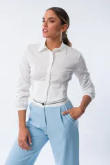 Camisa blanca de corte entallado con cuello clásico, cierre frontal de botones y mangas largas con puños elásticos. Presenta un diseño ajustado en la cintura con una banda elástica integrada que realza la silueta.