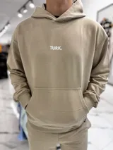 Conjunto de jogging beige que incluye un hoodie con capucha, bolsillo frontal tipo canguro y pequeño texto estampado en el pecho, acompañado de un pantalón tipo jogger con elástico en los tobillos.
