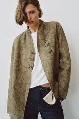 Blazer de corte recto confeccionado en tejido jacquard con diseño floral en tonos tostados. Presenta cuello mao, manga larga y cierre frontal mediante alamares decorativos. Incluye bolsillos de vivo en el delantero.