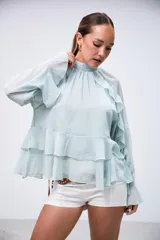 Blusa verde menta de gasa, con cuello alto fruncido, mangas largas amplias y detalles de volados en cascada en el frente y las mangas.