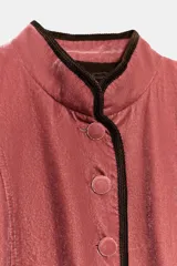 Blazer de terciopelo en color rosado con cuello subido y manga larga. Presenta cierre frontal con botones forrados y detalles de vivo en color oscuro a contraste en los bordes, cuello y puños. Incluye bolsillos delanteros de vivo.