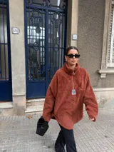 Buzo de corderito color marrón, con cuello alto y cierre frontal parcial. Presenta un diseño holgado con mangas largas y un parche decorativo en el frente.