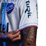 Camiseta de fútbol Umbro del Club Nacional de Football, modelo Home 2026. Diseño en color blanco con tejido jacquard que presenta un patrón de trofeos históricos en toda la prenda. Incluye cuello con detalles tricolores, logo de Umbro, escudo del club con estrellas y sponsors estampados en el frente y mangas.