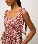 Blusa de viscosa y lino con estampado floral en tonos rojos y blancos. Presenta escote cuadrado, cuerpo ajustado con fruncido en la cintura y bajo con volado (peplum). Los breteles son anchos y se atan con lazos en los hombros.