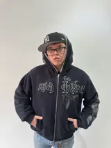 Campera reversible con cierre frontal, diseño oversize. Un lado es de algodón negro con estampados y bordados gráficos, mientras que el otro lado es de tela polar gris de pelo largo. Cuenta con capucha, puños y ruedo acanalados.