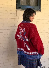 Saco tejido tipo jacquard color bordó con diseño floral en la espalda, cuello alto y cierre frontal de aluminio. Presenta detalles en azul y blanco en el cuello, puños y ruedo, con un patrón geométrico de rombos en la parte inferior.