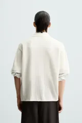 Camisa de hombre de corte relajado, color blanco roto, confeccionada en tejido de punto con estructura abierta y textura acanalada. Presenta cuello solapa y manga corta.