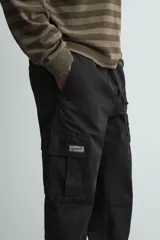 Pantalón cargo de corte slim fit en color negro, confeccionado en tejido de algodón con elasticidad. Presenta cintura elástica ajustable con cordón, bolsillos laterales tipo cargo con solapa y bajo acabado en puño elástico.