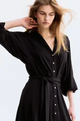 Vestido camisero midi de color negro, corte holgado con cuello, escote en V y cierre frontal de botones. Cuenta con mangas largas con puños anchos y un cinturón ajustable para anudar en la cintura.