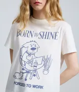 Remera de algodón color blanco con cuello redondo y mangas cortas. Presenta una estampa frontal en color azul con la frase 'Born to Shine' y una ilustración de un perro trabajando en una computadora.