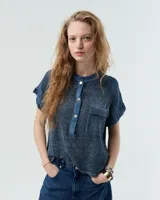 Remera blanca de lino con cuello mao, media cartera con broches metálicos y manga japonesa. Presenta un bolsillo frontal tipo plaqué, recortes laterales en sisa con detalles de encaje y ruedo redondeado con corte escalonado.