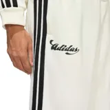 Pantalón deportivo Adidas House of Tiro color beige, con diseño de corte recto y ajuste holgado. Presenta las icónicas tres tiras laterales en color negro, logo de la marca y tipografía cursiva en el frente. Cuenta con cintura elástica con cordón ajustable.