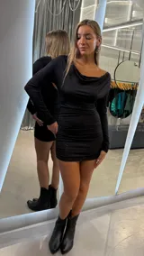 Vestido corto de manga larga en color negro con sutiles destellos brillantes. Presenta un escote drapeado tipo cowl y un diseño ajustado con frunces laterales que realzan la silueta.