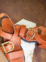 Sandalias de cuero marrón con tiras anchas cruzadas y hebilla metálica decorativa en forma de estribo. Poseen taco cuadrado de altura media y suela de goma negra.