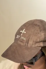 Gorra de gamuza color chocolate con bordado frontal en color crema que incluye una flor de lis y el texto 'EST. 1994'.