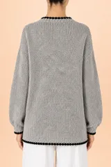 Cardigan de punto grueso en tono gris jaspeado, con cuello redondo y cierre frontal de botones. Presenta detalles de terminación en color negro con borde festoneado en el cuello, puños y ruedo, y mangas largas con puños ajustados.