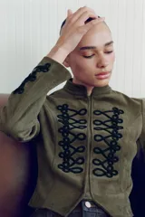 Chaqueta corta de antelina color verde militar, con cuello subido, manga larga y bajo asimétrico. Presenta detalles de alamares negros bordados en contraste en el frente y en los puños.