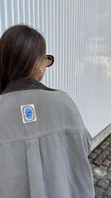 Sobrecamisa de denim en color gris con lavado desgastado, corte oversize y cuello de cuero sintético en color marrón chocolate. Cuenta con cierre frontal de botones metálicos.