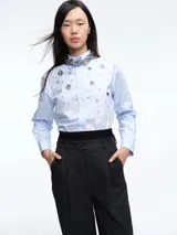 Camisa de algodón color celeste claro, con corte clásico, cuello camisero y cierre frontal de botones. Presenta apliques decorativos de pedrería y piezas metálicas circulares en la parte frontal.