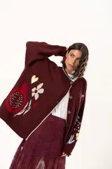 Saco de punto color vino con diseño holgado, cuello redondo y mangas largas. Presenta un gran bordado central en la espalda con forma de flor en tonos crema y rojo, acompañado de un pequeño bordado de sol y una frase bordada en la parte inferior.