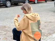 Campera tipo hoodie confeccionada en tejido de corderito color camel, con diseño de collage en la espalda que combina parches de tela en tonos verde oliva, naranja y estampados étnicos. Presenta un corte holgado y capucha integrada.