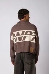 Campera tejida de punto con cierre frontal completo y cuello polo. Presenta un diseño de franja horizontal ancha en color crema con estampado gráfico oscuro sobre una base de color marrón. Posee puños y ruedo acanalados, con un pequeño parche de marca en el puño izquierdo.