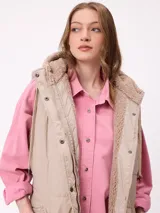 Chaleco largo acolchado color beige, con capucha forrada en sherpa, cierre frontal con cremallera y botones a presión, y bolsillos laterales.