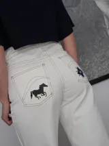 Pantalón vaquero de corte recto y tiro alto en color blanco marfil, con detalles de costuras en contraste y estampa decorativa en la parte inferior de las piernas, que incluye figuras de caballos y patrones geométricos. Presenta aberturas laterales en los tobillos.