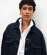 Campera tipo overshirt de denim en color azul oscuro, con corte amplio y largo extendido. Presenta cuello clásico, cierre frontal con botones metálicos, mangas largas y dos bolsillos delanteros con solapa.