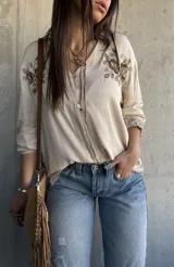 Camisa beige de manga larga con cuello en V, cierre frontal de botones y delicados bordados florales en marrón en la zona del pecho y en los puños.