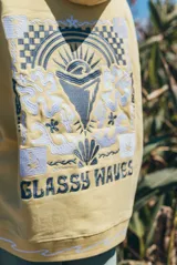 Hoodie amarillo de corte holgado con capucha, con un diseño bordado en la espalda que incluye motivos de olas, sol y la inscripción 'Glassy Waves'.