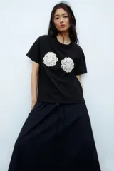 Remera negra de punto de algodón, de corte holgado con hombros caídos, cuello redondo acanalado y mangas cortas. Presenta dos apliques decorativos circulares de crochet en tono crudo a la altura del pecho.