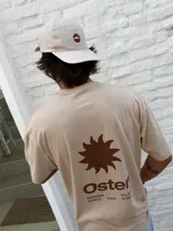 Remera oversize color beige con cuello redondo y manga corta. Presenta un estampado minimalista en color marrón en el pecho, que incluye un símbolo similar a un sol o estrella y texto.