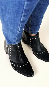 Botín corto de cuero negro con textura combinada en la puntera y el talón. Cuenta con punta fina, apliques metálicos en forma de tachas y cierre lateral plateado.