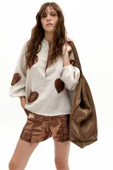 Camisa oversize de lino color beige, con cuello mao, cierre frontal con botones de nácar y mangas abullonadas con puño elástico. Presenta apliques de corazones en material metalizado tono bronce.