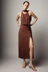 Vestido midi de color marrón con cuello halter y lazada en la nuca. Presenta un diseño con espalda descubierta, frunces en la cintura y una abertura lateral en la falda.