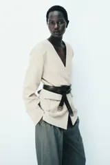 Blazer de punto grueso en color beige, con diseño sin cuello, escote en V, hombreras y cintura entallada. Cuenta con cierre de un botón frontal y bolsillos delanteros con solapa.