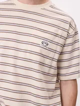 Remera de corte holgado con diseño de rayas horizontales en tonos beige, marrón y azul claro. Presenta cuello redondo, mangas cortas y un bolsillo frontal en el pecho con el logo de la marca bordado.