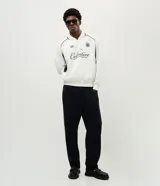 Buzo deportivo de felpa en color blanco con detalles en negro. Presenta cuello tipo polo, mangas largas con puños acanalados y diseño con recortes laterales. Incluye estampados gráficos en el frente con tipografía cursiva y emblema circular.