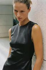 Vestido corto de cuero sintético negro con cuello redondo, manga sisa, hombreras y costuras marcadas que aportan estructura. Presenta bajo con volumen y cierre en la espalda con cremallera metálica.