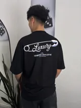 Remera negra de corte oversize con estampado frontal de estilo barroco/urbano que incluye un ángel con alas, cadenas, una cruz y la inscripción "Luxury".