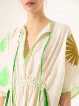 Túnica midi de color blanco crudo, con mangas cortas amplias, escote en V con cordones verdes ajustables y cintura fruncida. Presenta detalles de bordados geométricos verdes y soles marrones en las mangas y el ruedo.