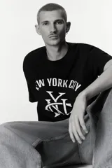 Remera de algodón color negro con cuello redondo acanalado, mangas cortas y hombros caídos. Presenta un estampado frontal en color blanco con la inscripción New York City y un monograma central.