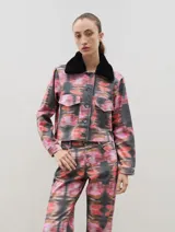 Chaqueta de denim con estampado abstracto en tonos rosados, naranjas y negros. Presenta un corte recto, cierre frontal con botones metálicos, bolsillos frontales con solapa y un cuello de piel sintética desmontable en color negro.