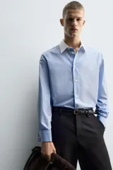 Camisa de corte regular fit confeccionada en algodón, con estampado de rayas verticales en tonos azul y blanco. Presenta cuello italiano en color blanco a contraste, cierre frontal de botones y mangas largas con puños dobles.