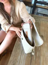 Cartera tipo bandolera de cuero color crudo, con diseño rectangular, solapa frontal con detalle de costura decorativa en el borde y correa ajustable.