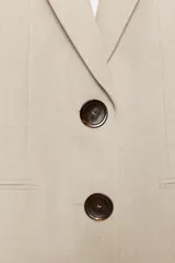 Blazer sastre de corte recto, color beige, con cuello solapa, hombreras marcadas y cierre frontal de dos botones oscuros. Cuenta con bolsillos con solapa en el frente y un bolsillo ojal en el pecho.