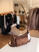 Cartera tipo baguette con estampado animal print, confeccionada en material suave símil gamuza. Presenta un bolsillo frontal con solapa y cierre, compartimento principal con cierre superior, y asa larga regulable de cuero sintético marrón.